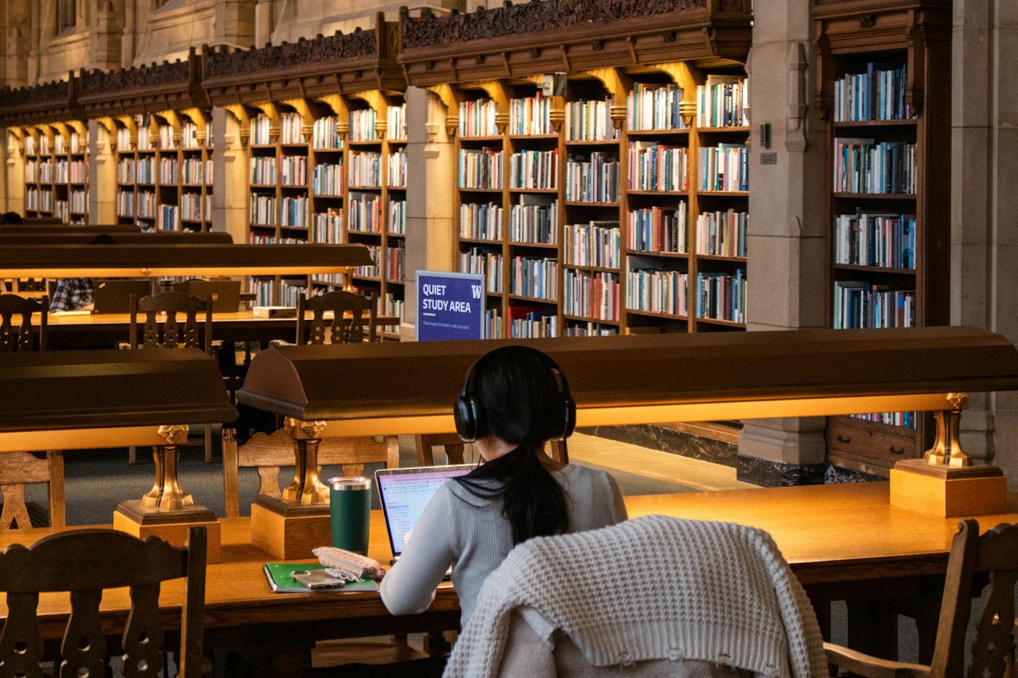Eine Person mit Kopfhörern arbeitet an einem Laptop an einem Holztisch in einer Bibliothek.